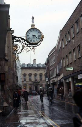 Pedestrians walking down a street with a clock overhead