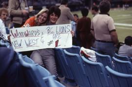 Two unknown woman sitting in the stands of BC Place holding a sign that reads: "Woodward's N...