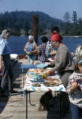 Woman with a red head scarf taking food from a buffet table surrounded by other people milling about