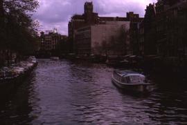 River with a boat in the foreground and buildings on the right and cars and trees on the left of ...