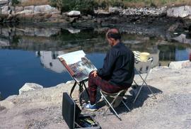 Unknown man with his back to the camera painting on a rocky shoreline with water in front of him