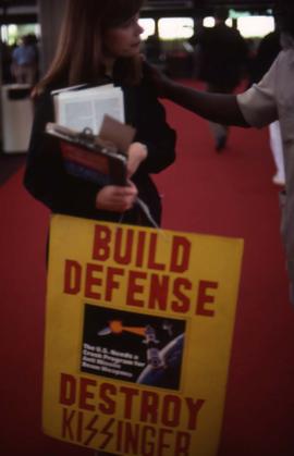 Woman holding several clip boards and a sign that reads: "Build Defence Destroy Kissinger"