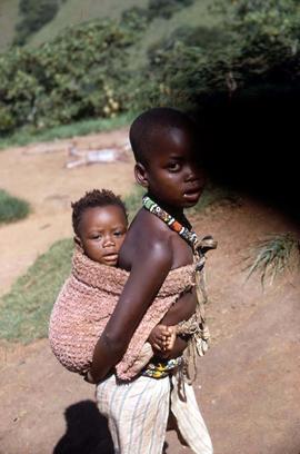 Unidentified South African boy carrying an unidentified infant on his back, wearing traditional e...