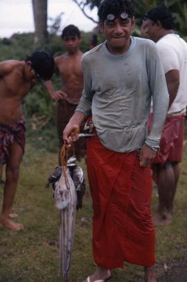 Four fishermen, one standing in front of them holding up fish and an octopus