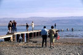 Unidentified groups of people standing on a pier, as well as swimming in a body of water